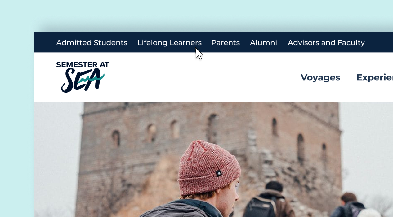 Person wearing a pink beanie stands in front of a historic stone building with other visitors.